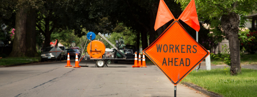 Orange and Black "Workers Ahead" Sign in Residential Neighborhood with Equipment in Background, Daytime
