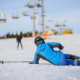 Young woman skier in blue ski suit after the fall on mountain slope trying get up against ski-lift. Ski resort. Winter sports concept.