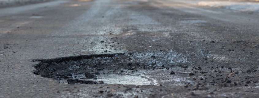 A pothole in the road and a car in the background that is moving towards this pothole