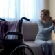 Disabled lady sitting on sofa and looking in window, empty wheelchair in room